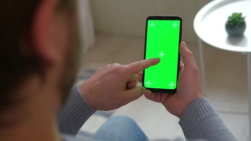 Young Man Sitting at Home Holding Smartphone Green Mockup Screen in Hand Male Person Using Chroma