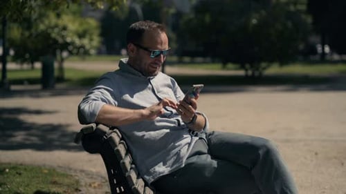 Hombre enviando mensajes de texto en un teléfono inteligente sentado en un banco en City Park