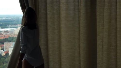 Young woman unveiling curtains admiring city view in morning light super slow motion