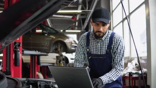 Experienced Mechanic in Uniform is Using a Laptop While Repairing Car in Auto Service