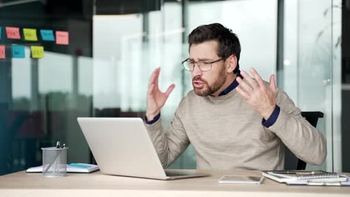 Angry businessman arguing during video call on laptop while sitting at desk in office.