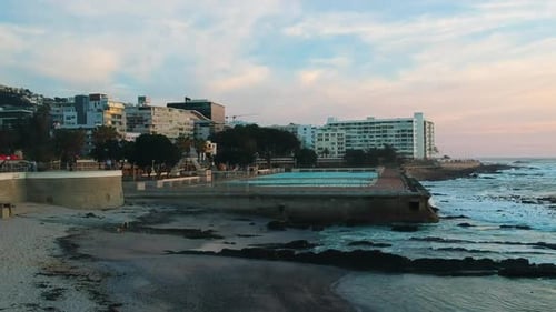 A drone slowly ascending above a beach in Sea Point, Cape Town during sunset with bathing pools in t