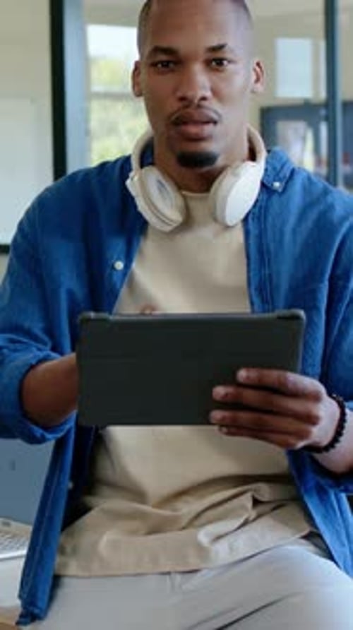 Vidéo verticale : Homme souriant en chemise bleue tenant une tablette dans une pièce lumineuse, riant du contenu