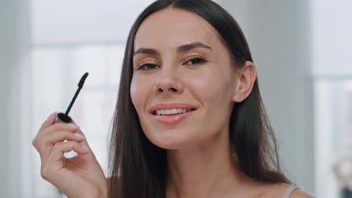 Smiling woman applying mascara to eyelashes indoors