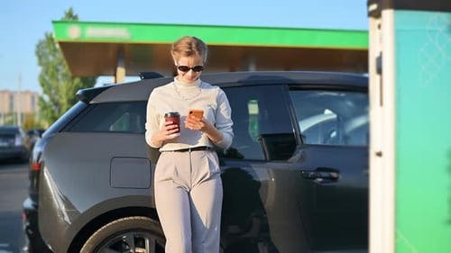 A young blonde woman with smartphone and coffee at a car charging station with electric car nearby i