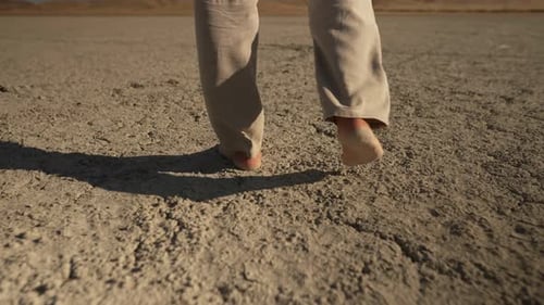 A Man in Summer Trousers Walks Along the Bottom of a Dry Lake Stepping on Dry Soil with Bare Feet