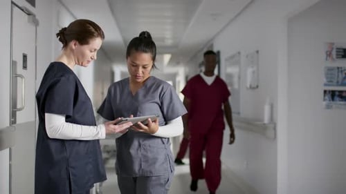Diverse female doctors discussing work, using tablet in corridor at hospital, slow motion