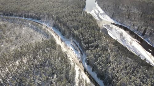 Aerial view of a winding river through a dense snow-covered forest in winter, with a curving road ru