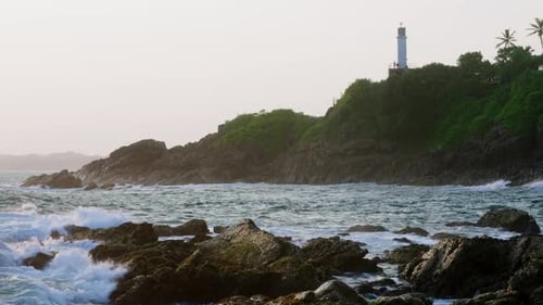 Waves Crash Against Rocks at Sunset with Lighthouse on a Hill Video Captures Ocean Navigation Safety