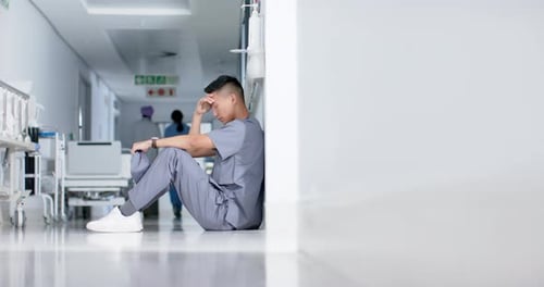 Stressed asian male surgeon sitting and leaning on wall in hospital in slow motion
