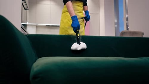 Close Up in an Apartment Cleaner in a Yellow Apron Does a Steam Cleaning of a Green Sofa with