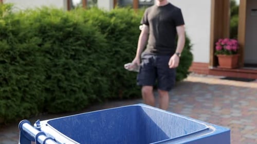 Unrecognizable man walks out to throw plastic bottles into trash bin in garden