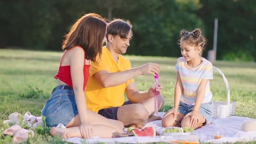 Family Enjoys a Sunny Afternoon in the Park Making Bubbles and Having a Picnic with Fresh Fruits and