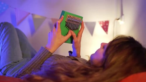 Female Musician Relaxing on Bed Gently Strumming Green Kalimba Colorful Pennants Hanging Warm