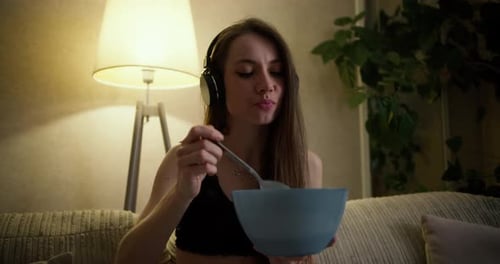 Woman Listening to Music in Wireless Headphones While Eating Lunch Sitting on Sofa in Dark Cozy