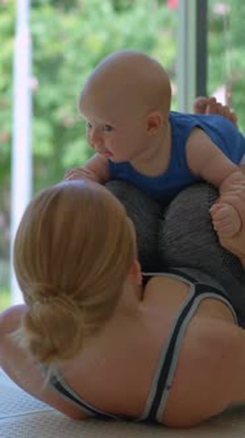 Joven madre hace ejercicio con su hijo de 3 meses en casa junto a una ventana grande con un exuberante paisaje tropical