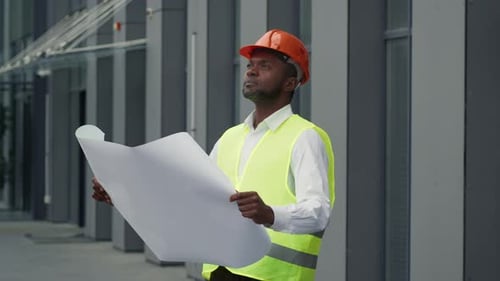 Arquitecto negro profesional con casco y chaleco protectores que estudia un plano en papel y
