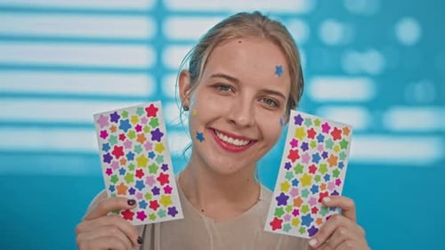 Positive Young Woman with Colorful STickers in Hands and on Face against Blue