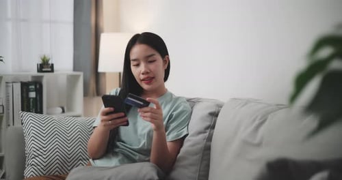Young woman uses phone and credit card to shop online on sofa at home.