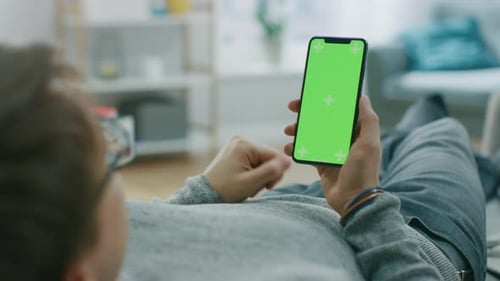 Man at Home Lying on a Couch using Smartphone with Green Mock-up Screen, Doing Swiping, Scrolling G