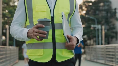 Man Builder with Laptop and Coffee Walks Along Bridge Against Green Park Closeup