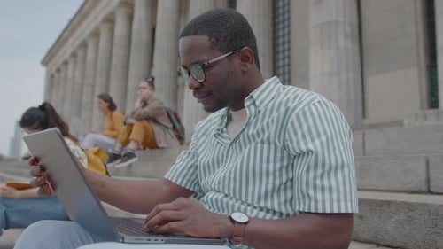 Black Student Typing on Laptop on Stairs Outside the University