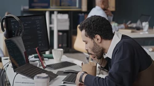 Middle Eastern Man Eating Lunch at Desk and Coding on Laptop in IT Office