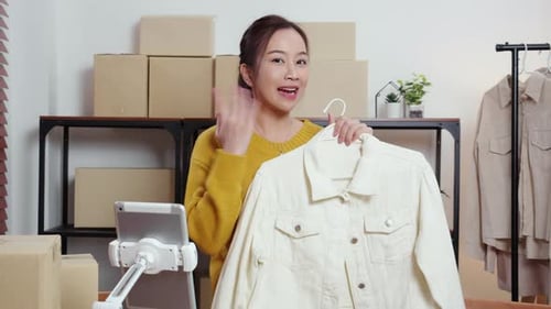 Two young asian woman live online clothing store while showing clothing with tablet on social.