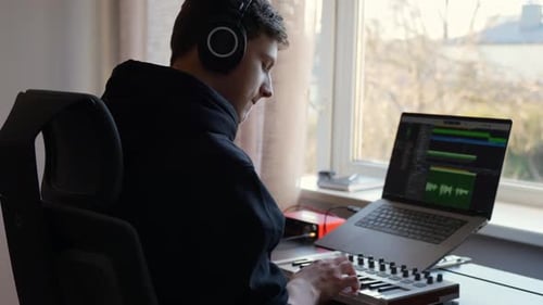 Young man producing music in home studio, wearing headphones and actively composing digital track