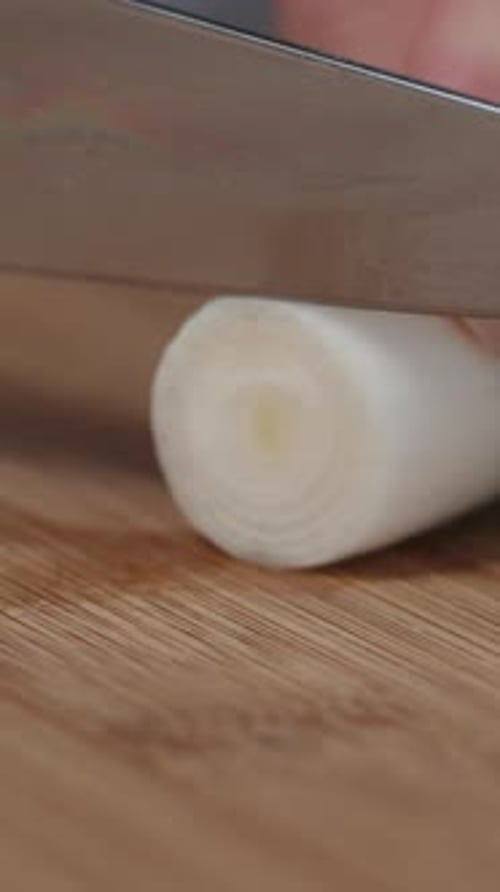 Vertical video. A woman cuts off the roots and slices the stem of a leek on a cutting board