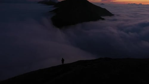A tilt up drone shot of a man standing by himself on the edge of a mountain overlooking a bed of clo