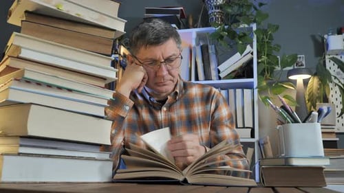 Man Reading Book at Desk Surrounded by Stacks