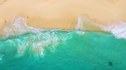 Aerial drone footage of massive turquoise waves crashing on a sandy tropical beach.