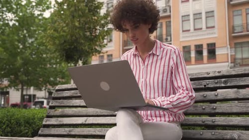 Smiling hipster woman student works on laptop on city street bench outdoors