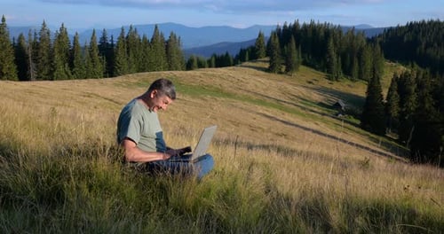 A Man Sits on a Beautiful Meadow in the Mountains Works on a Laptop