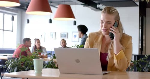 Happy caucasian casual businesswoman talking on smartphone using laptop in office, slow motion