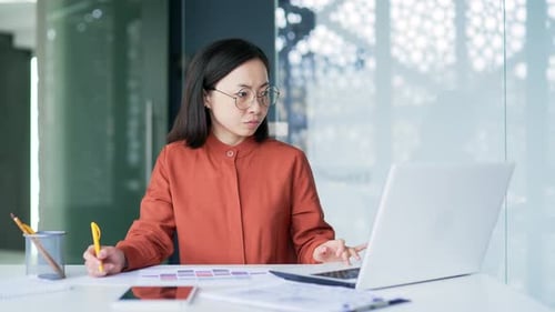 Busy asian businesswoman financier making financial calculations using typing on laptop, taking note