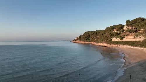 Calm waves hit the shore of a lonely beach, where a mild hill is rising above the water surface