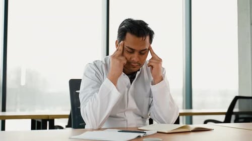 Stressed Frustrated Indian Doctor with Headache Sitting Alone at Work Desk in Hospital Sad Young Gp