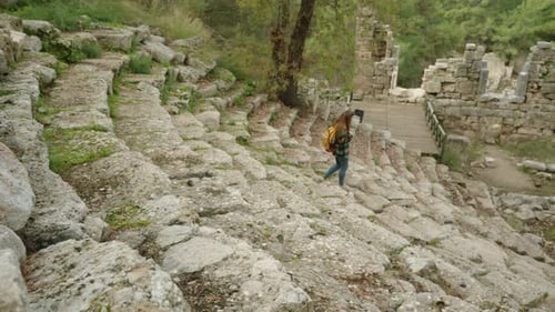 A female tourist with a backpack descends the steps of the ancient Greek amphitheater and sits.