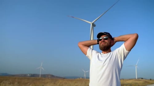 Hombre barbudo feliz y relajado levantando los brazos disfrutando de un viaje por la naturaleza, puesta de sol en el campo, molino de viento