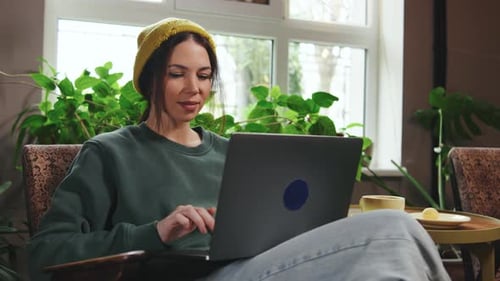 Woman Enjoying Coffee in a Cozy Cafe While Working on a Laptop Surrounded By Plants and Natural