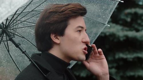 Young Man in a Coat Close Up Walking Under a Transparent Umbrella in the Autumn City Park and