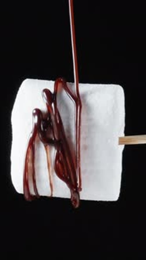 Vertical. White marshmallows on a stick. It rotates and a stream of chocolate pours on it. Close-up