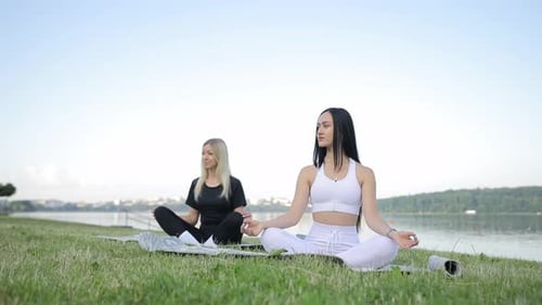 Two Young Women Practice Yoga While Sitting on a Mat By the Lake