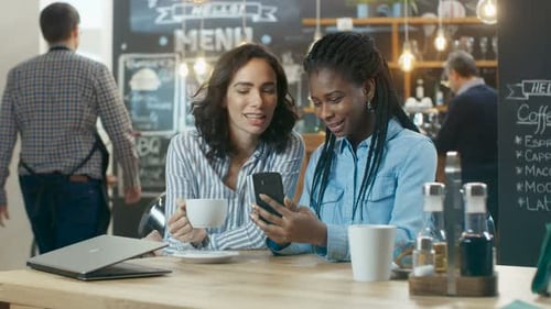 Two Beautiful Girlfriends Sit in the Coffee Shop and Chat, One Shows Something on Her Smartphone, t