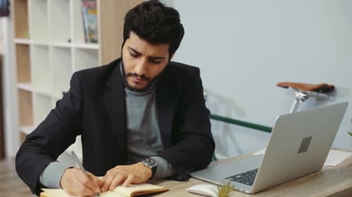 Focused Man Writing Notes at Desk with Laptop