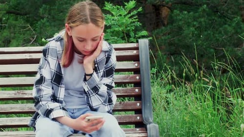 Sad Boring Teen Girl Sitting on Bench in Park Browsing Smartphone in Summer Day
