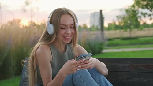 Lady in Headphones Listens to Music Sitting on Bench in Park