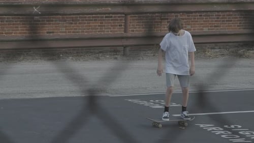 Teen skater on a skate board skating a line in a back alley parking lot as seen through a fence.